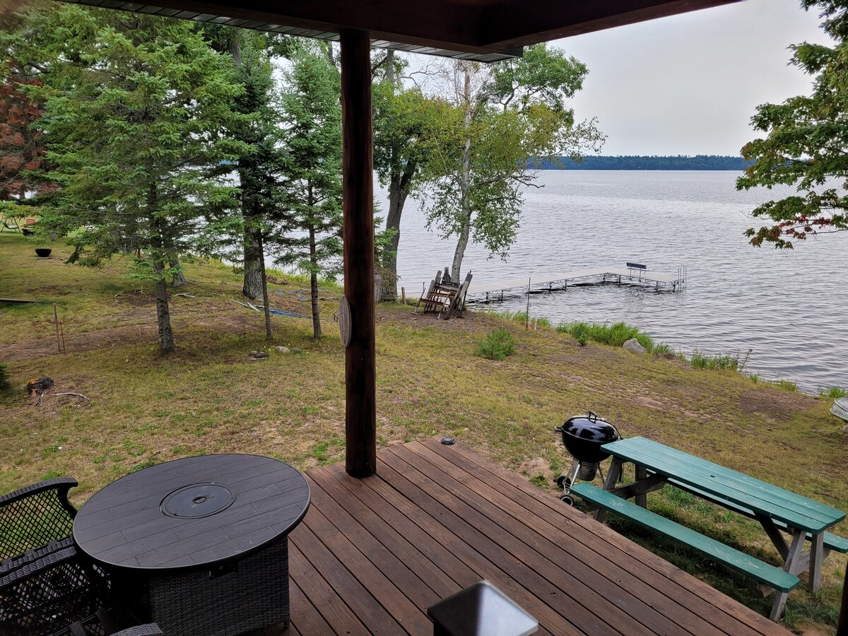 A wooden deck extends towards a tranquil lake, framed by lush trees. A round table and chairs are positioned nearby, with a charcoal grill visible to the side. A small dock is accessible from the grassy area leading to the water's edge.