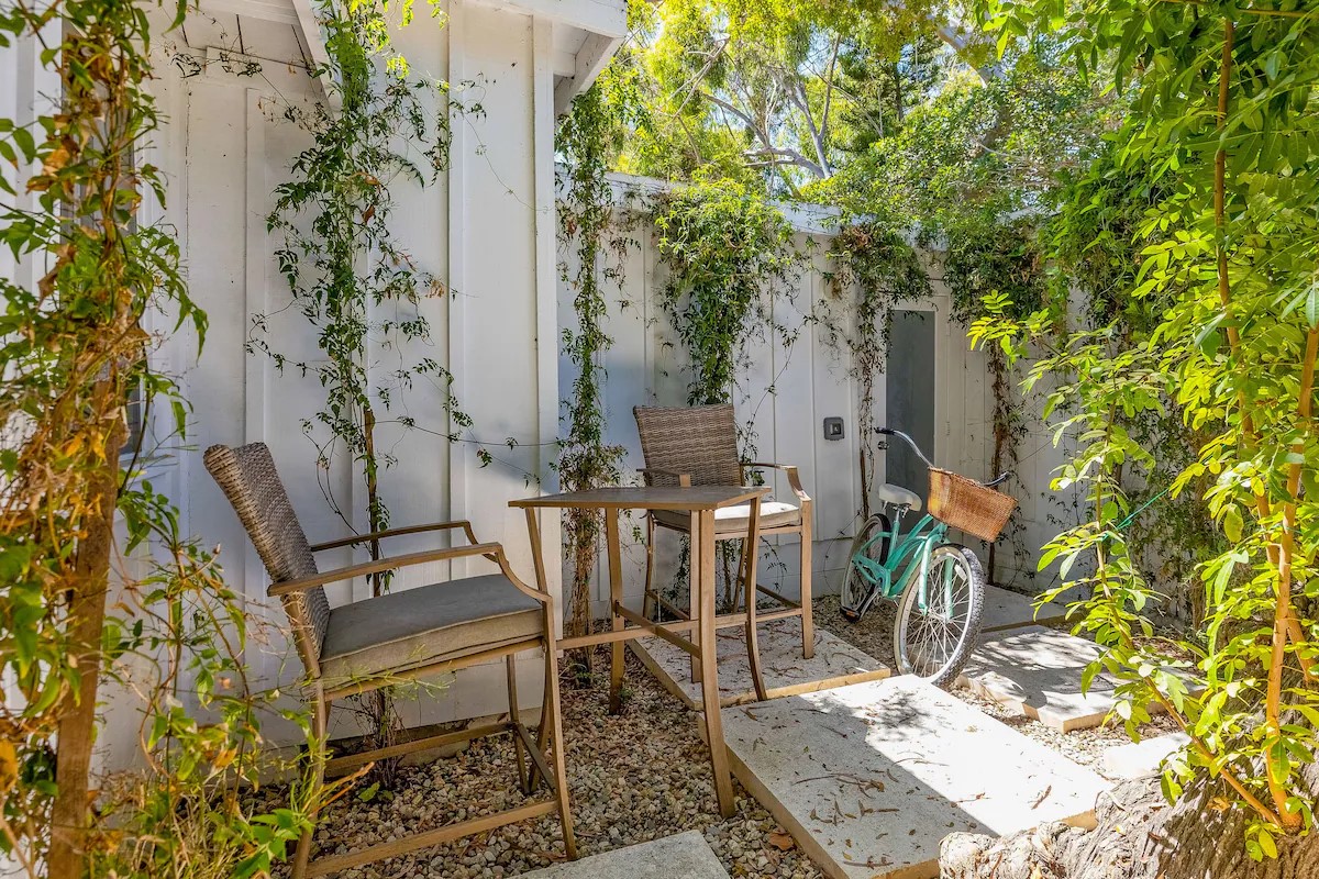 An outdoor patio area features two wooden chairs with soft cushions positioned beside a small table. A vintage bicycle rests against the wall, surrounded by greenery that adds a refreshing touch. Sunlight filters through the leaves, creating a serene environment.
