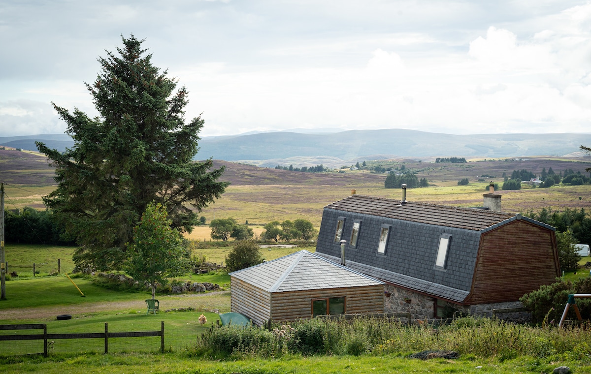 A charming holiday apartment is nestled among the highlands, surrounded by lush green fields and expansive landscapes. The wooden and stone structure features a sloped roof with skylights, offering scenic views of the rolling hills and distant mountains.