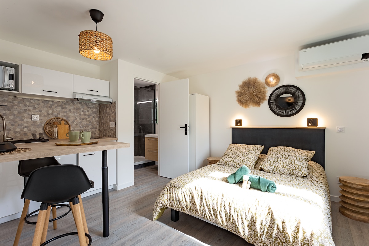 A functional studio is presented with a cozy sleeping area and a kitchenette. The bed features a patterned duvet, complemented by bedside lighting. A small dining area includes two stools at a counter. A door leads to a modern bathroom, visible in the background.
