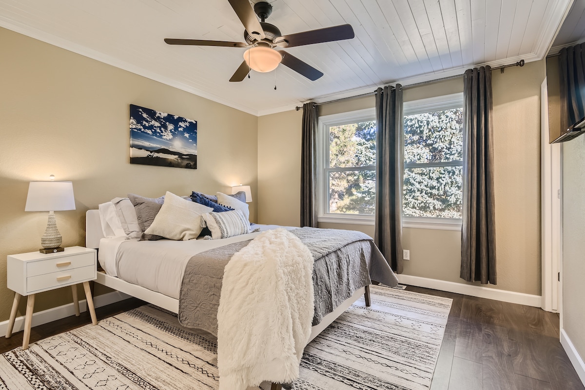 A serene bedroom features a spacious bed with neutral linens, layered with a soft throw blanket. Two windows allow natural light to brighten the room, complemented by modern curtains. A ceiling fan adds a practical touch, while a small side table and artwork enhance the calming atmosphere.