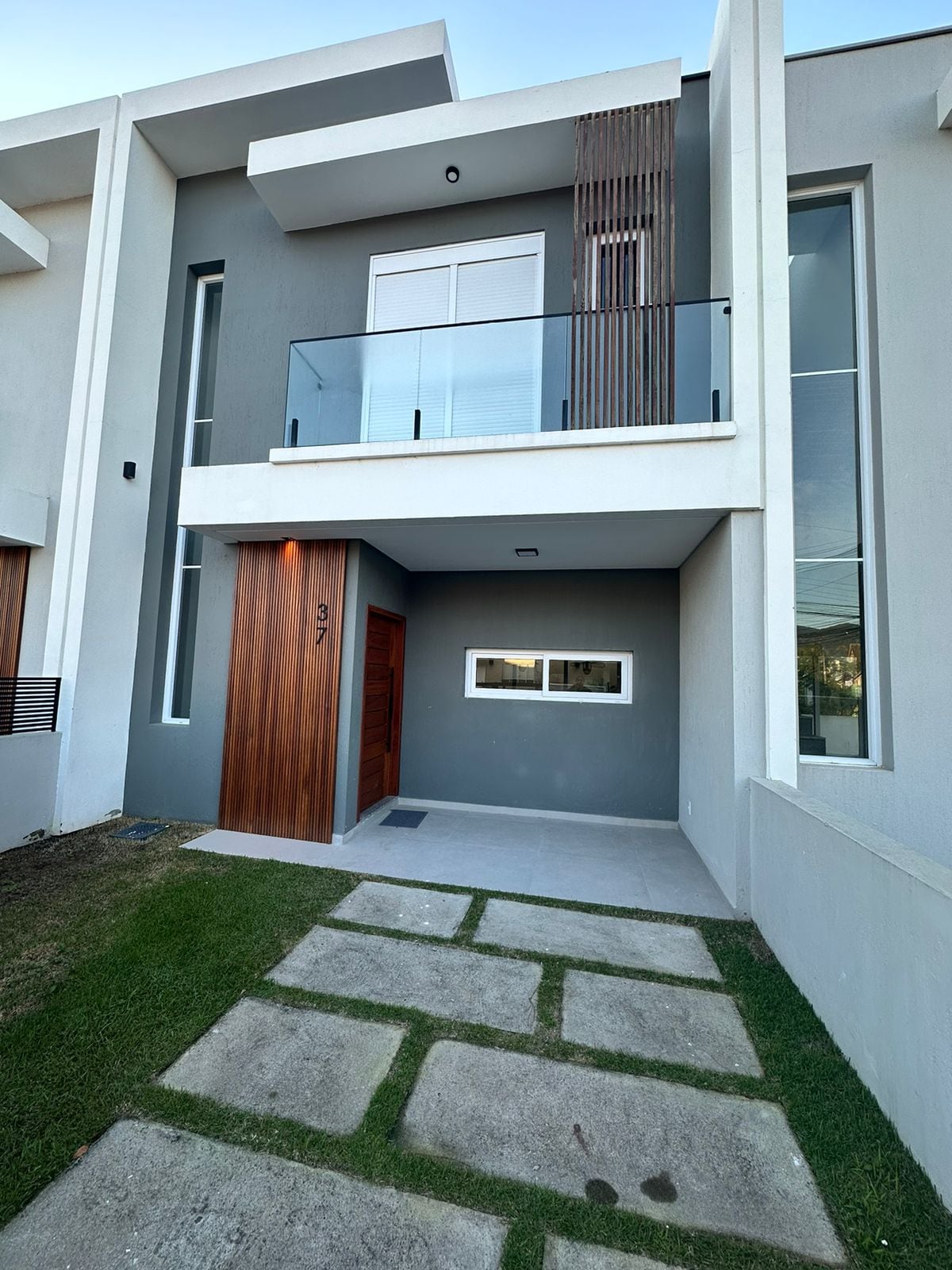The modern exterior features a sleek design with a combination of grey walls and natural wood accents. A spacious front pathway leads to a welcoming entry door, with a glass balcony above. Green grass borders the stone walkway, enhancing the overall aesthetic.