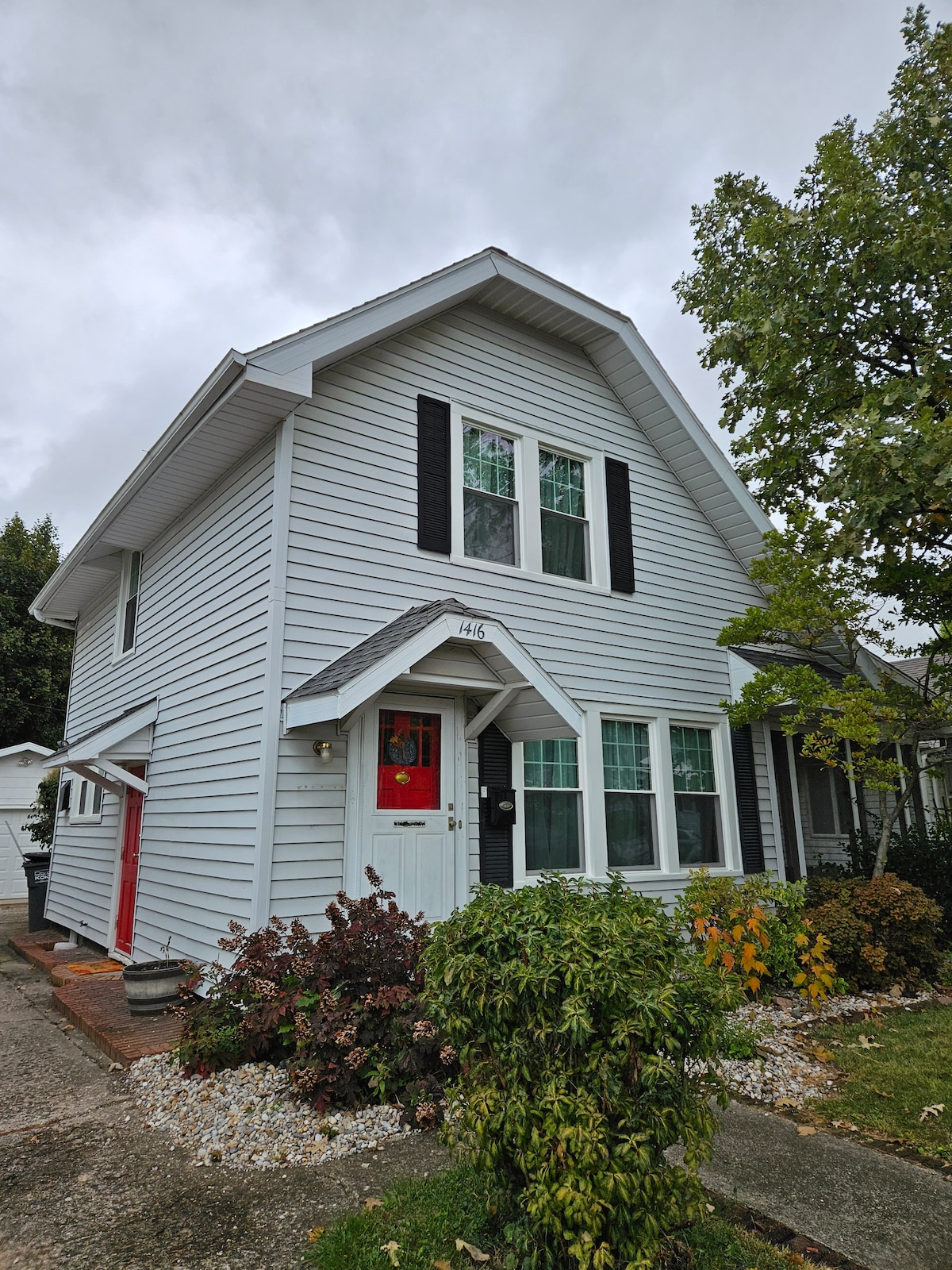A two-story home is shown with a light gray exterior and black shutters. The front porch features a welcoming red door. Lush greenery and colorful plants surround the entrance, providing a vibrant contrast against the home’s facade.