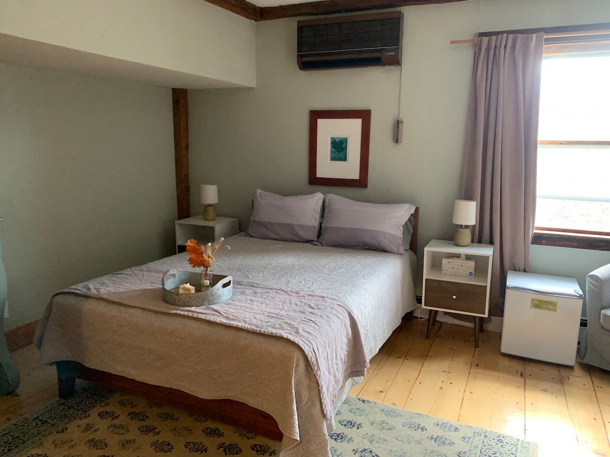 A spacious guest room features a queen size bed with a light-colored quilt. A bedside table is on each side, holding modern lamps. A window allows natural light to enter, complemented by soft curtains. The wooden flooring adds warmth, and a small dresser is visible in the corner.