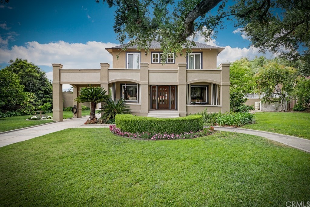 A grand two-story home with a light-colored exterior features arched windows and a double-door entrance. Lush green lawns surround the house, complemented by manicured flower beds and tall palm trees. A large oak tree provides shade nearby, enhancing the inviting curb appeal.