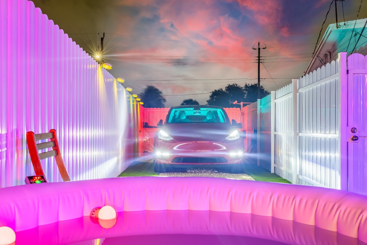 A Tesla Model Y is parked in a well-lit driveway, bordered by a white fence. The scene is enhanced by colorful lighting, while a hot tub with pink accents sits in the foreground. A tranquil evening sky adds to the inviting ambiance.