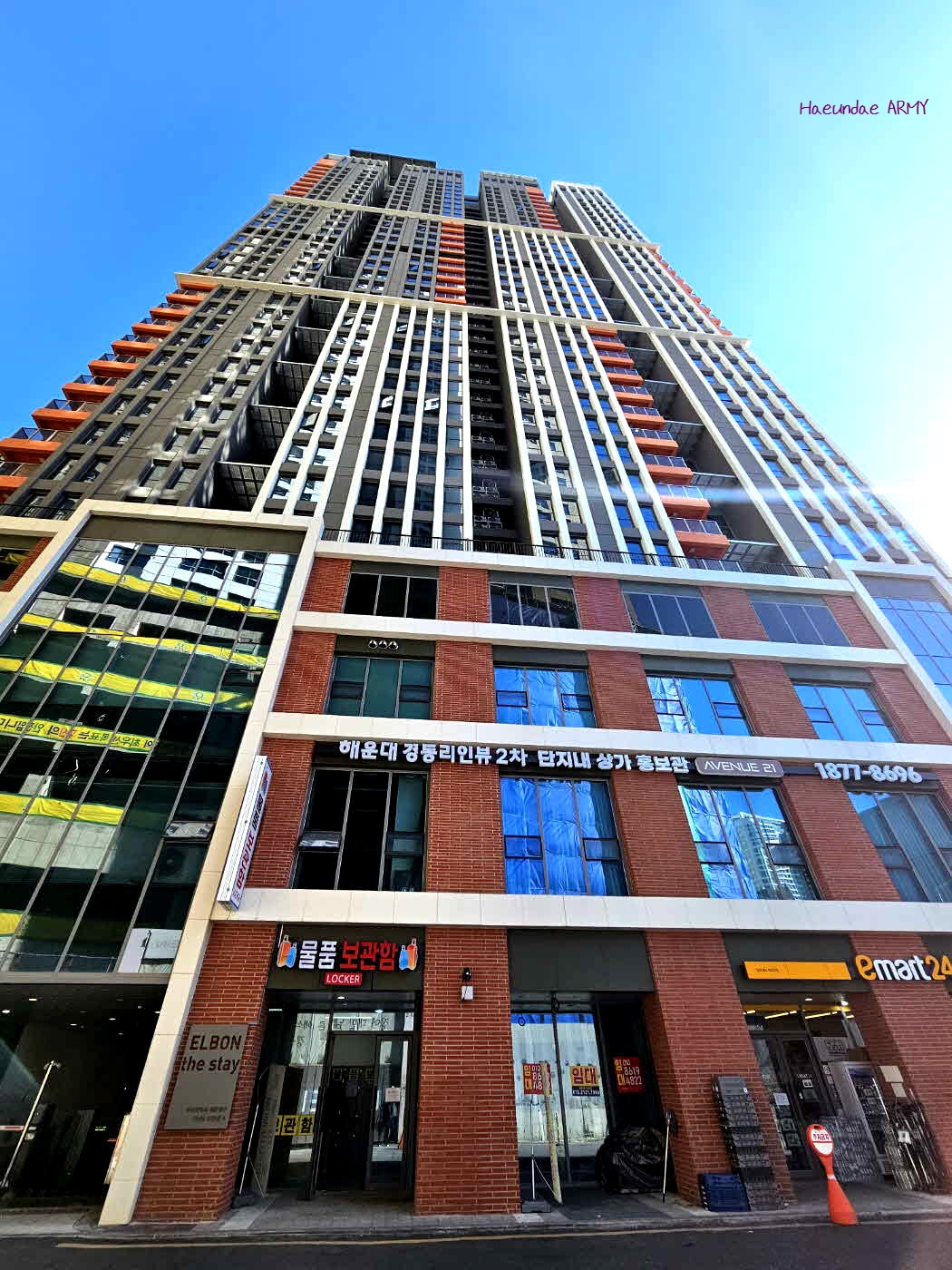 The image showcases a modern high-rise building with a distinctive architectural design. The structure features a mix of glass and brick facades, emphasizing vertical lines. A clear blue sky provides a bright backdrop, while the entrance is marked by signs for businesses located within.