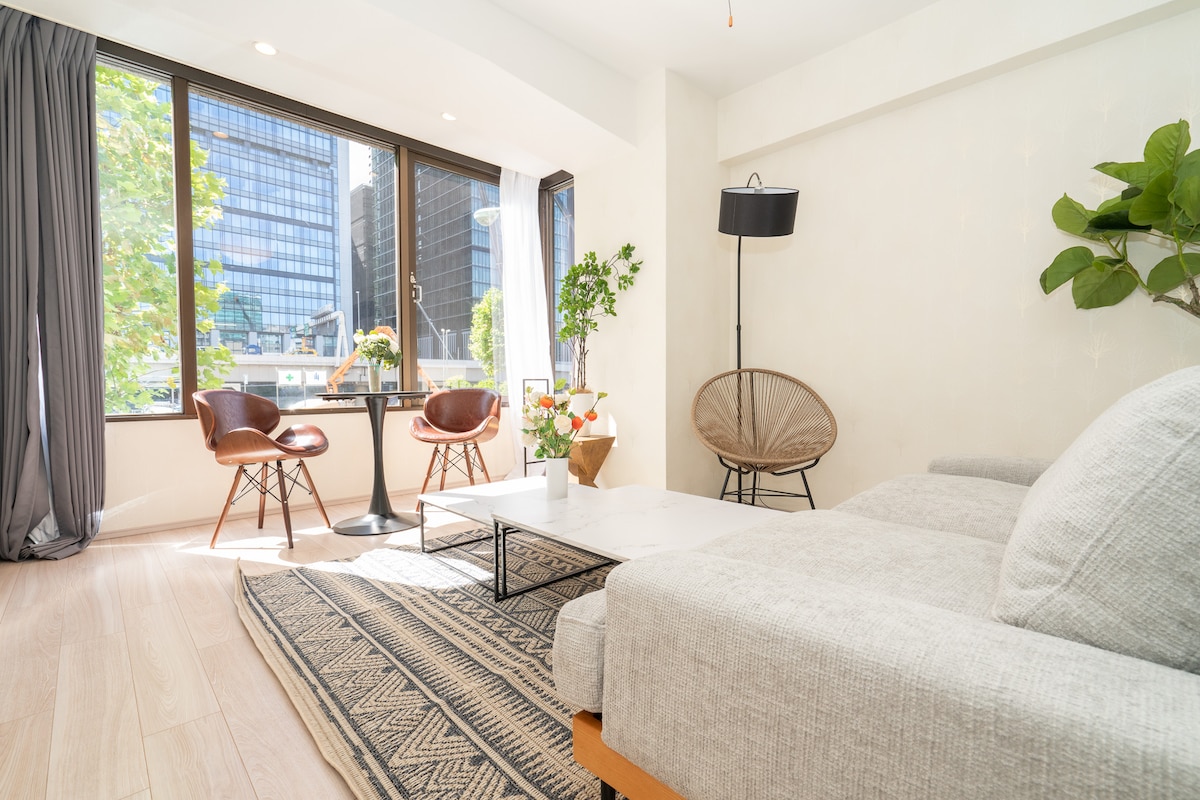 A bright living area features large floor-to-ceiling windows allowing natural light to fill the space. A comfortable sofa sits beside a decorative rug, while a coffee table is surrounded by two modern chairs and a small dining table, offering a cozy gathering spot.
