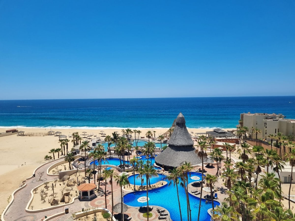 An expansive view showcases a shimmering blue pool surrounded by palm trees, leading to a sandy beach and the ocean beyond. A unique thatched-roof structure adds character to the landscape, with lounge chairs scattered poolside, inviting relaxation under the clear sky.