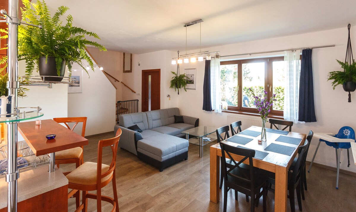 An open living and dining area features a spacious gray sectional sofa facing a glass coffee table. A wooden dining table with black chairs is surrounded by natural light from large windows. Potted plants add greenery to the room, enhancing the inviting atmosphere.