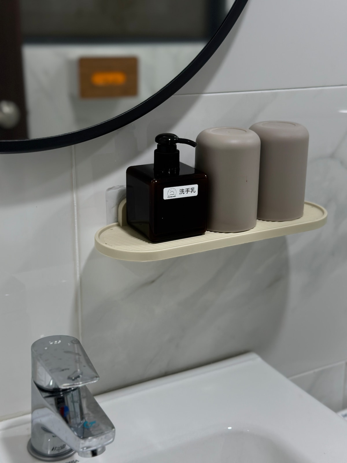 A modern bathroom shelf is shown, featuring two cylindrical grey containers and a black pump bottle with a label. The shelf is positioned above a sleek faucet, and a round mirror reflects the elements within the space.