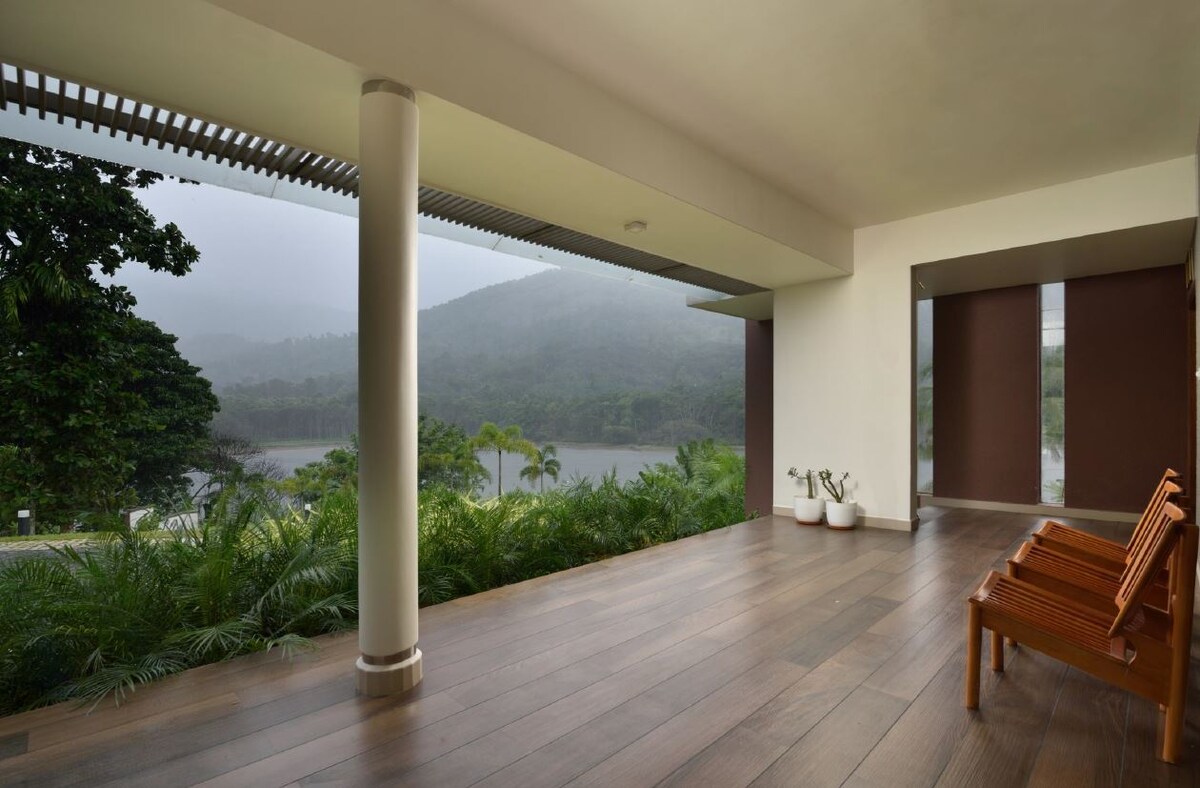 A spacious outdoor sitting area features two wooden chairs, offering views of lush greenery and a misty landscape. Large windows and sliding doors provide a seamless connection to the surrounding nature, enhancing the tranquil atmosphere of the space.