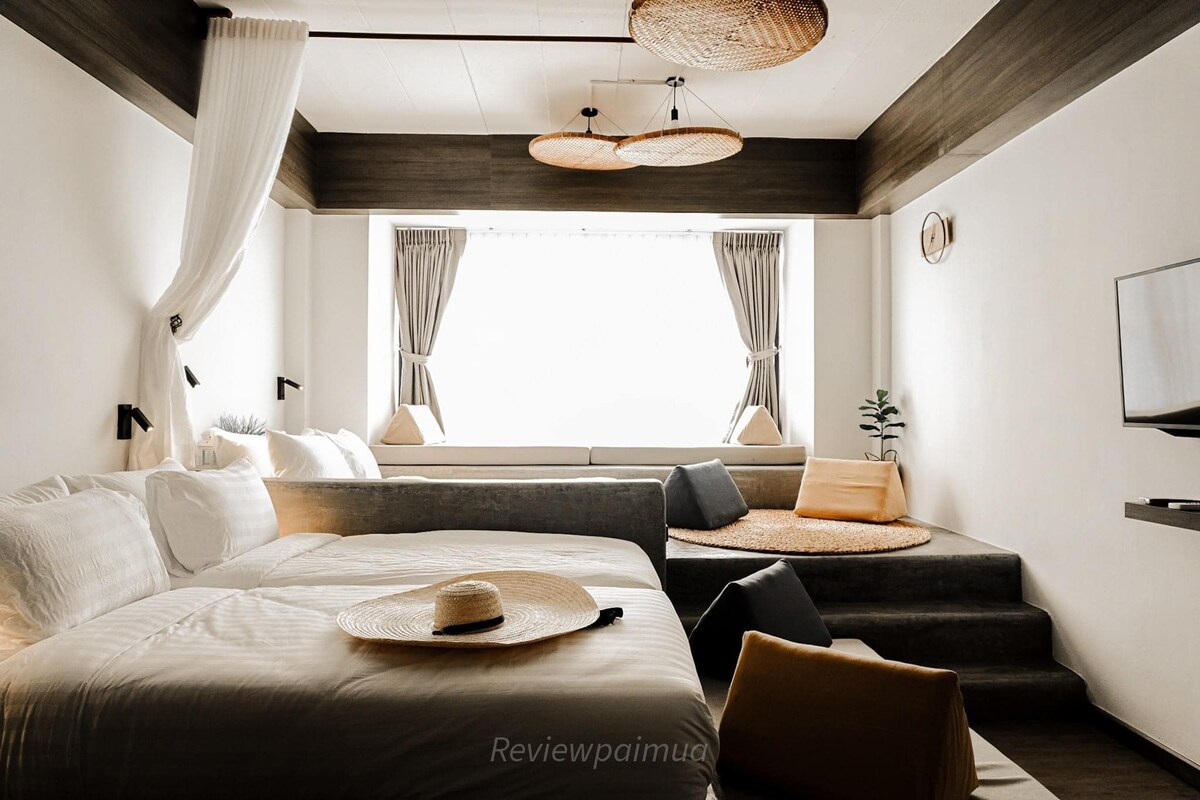 A spacious room features a large bed and a cozy seating area with cushions and a round mat. Natural light filters through large windows with soft curtains. Decorative woven light fixtures hang from the ceiling, complementing the neutral tones of the decor.