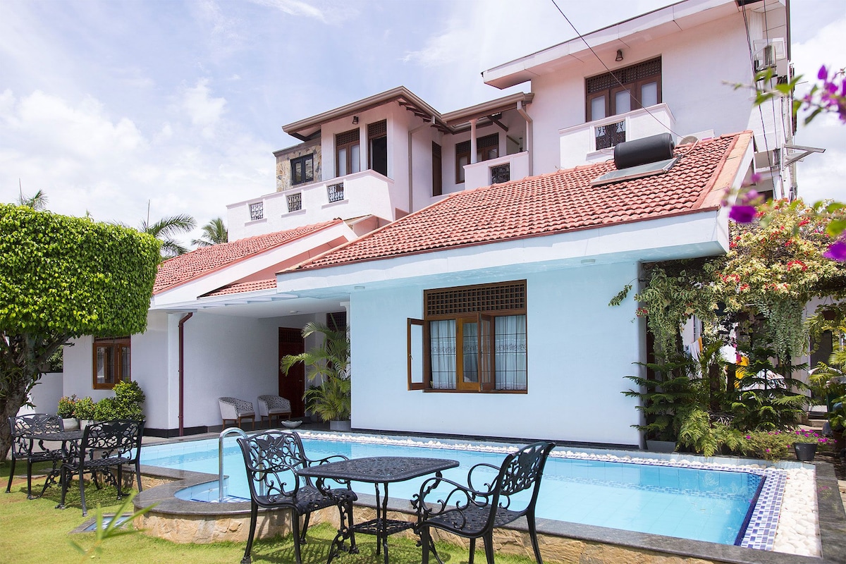 A charming villa is presented with a well-maintained garden surrounding a pool. Outdoor seating arrangements consist of wrought iron tables and chairs, inviting relaxation. The house features a terracotta roof and large windows, allowing natural light to enhance the tranquil environment.