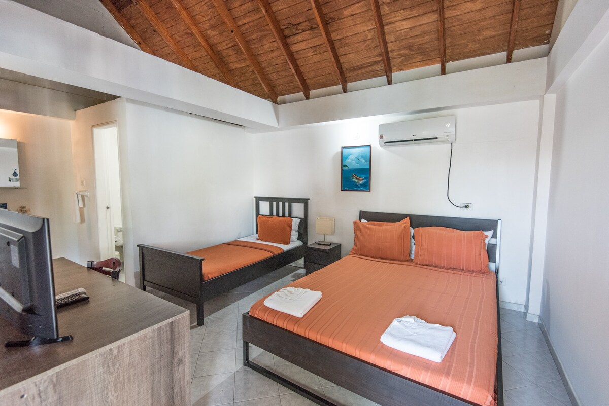 A cozy bedroom features a spacious bed along with a separate single bed, both dressed in orange linens. Light wood accents are complemented by a flat-screen TV and a nightstand with a lamp. A piece of artwork depicting ocean scenery is displayed on the wall.