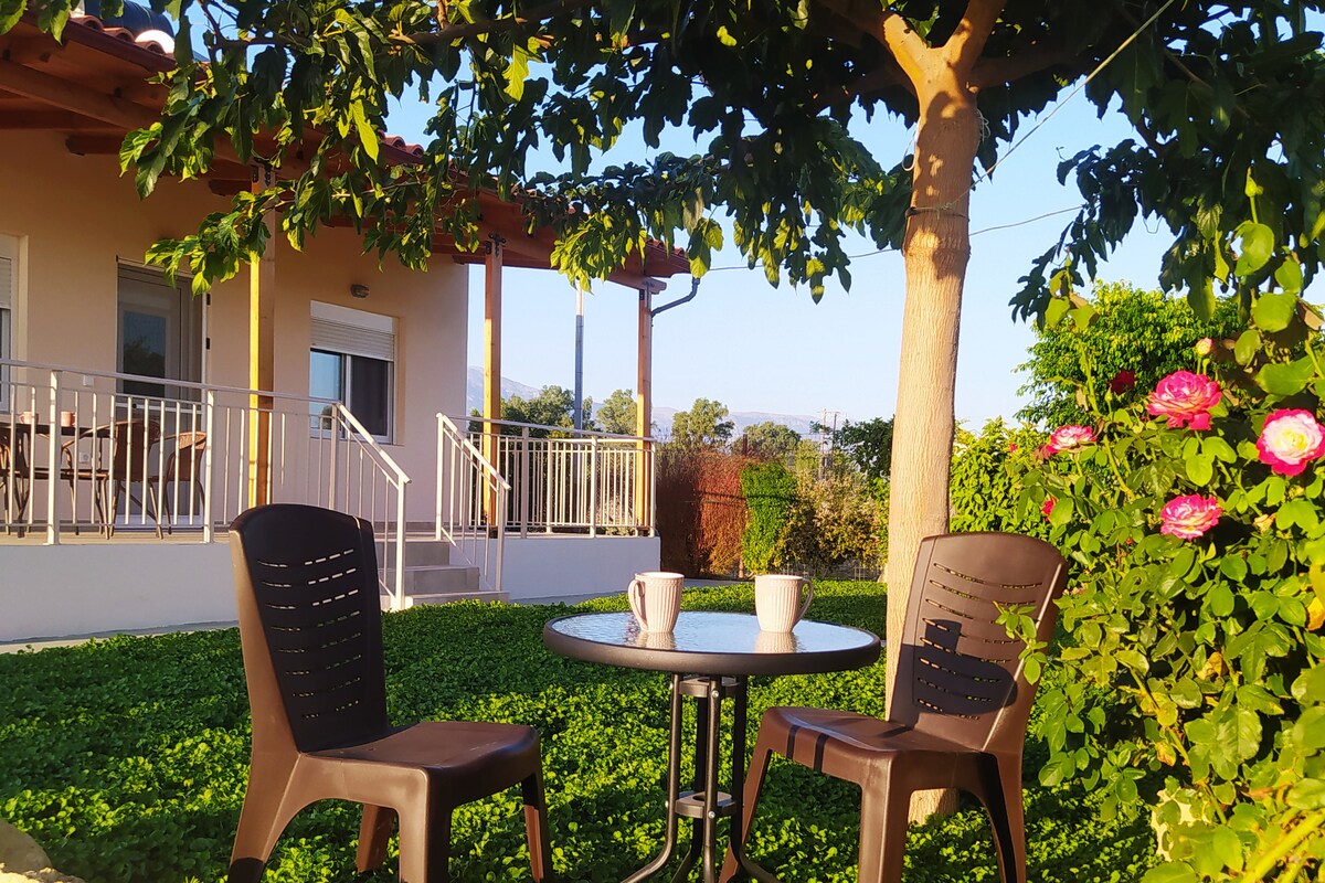 A shaded outdoor seating area is set with two chairs and a small table, positioned amid lush greenery. The house, featuring a covered porch, is visible in the background, surrounded by vibrant flowers and trees under clear skies.