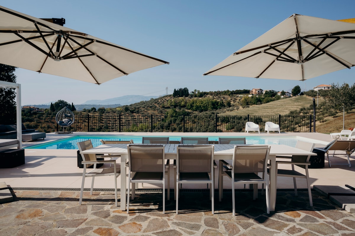 An outdoor dining area is presented with a large table and multiple chairs beneath two shaded umbrellas. In the background, a private swimming pool reflects the surrounding landscape, with rolling hills and distant trees enhancing the space.