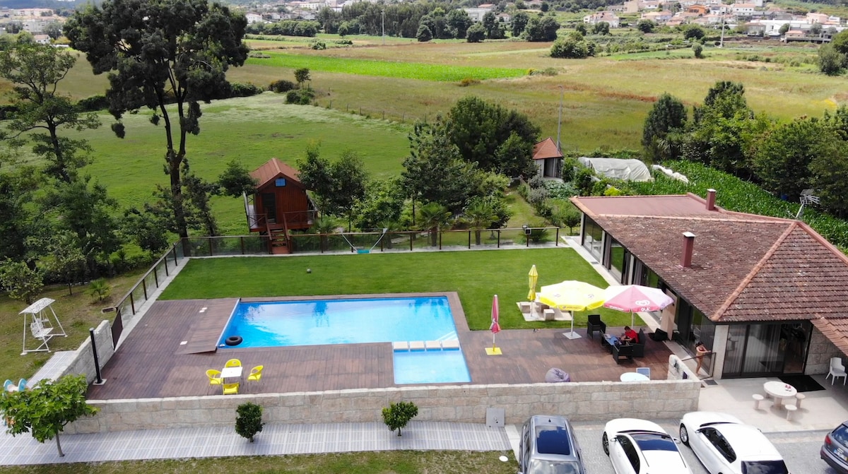 An aerial view showcases a large outdoor pool surrounded by an expansive wooden deck. Colorful lounge chairs and umbrellas are arranged for relaxation, while lush green fields and trees provide a natural backdrop. A well-kept garden and seating area are also visible.
