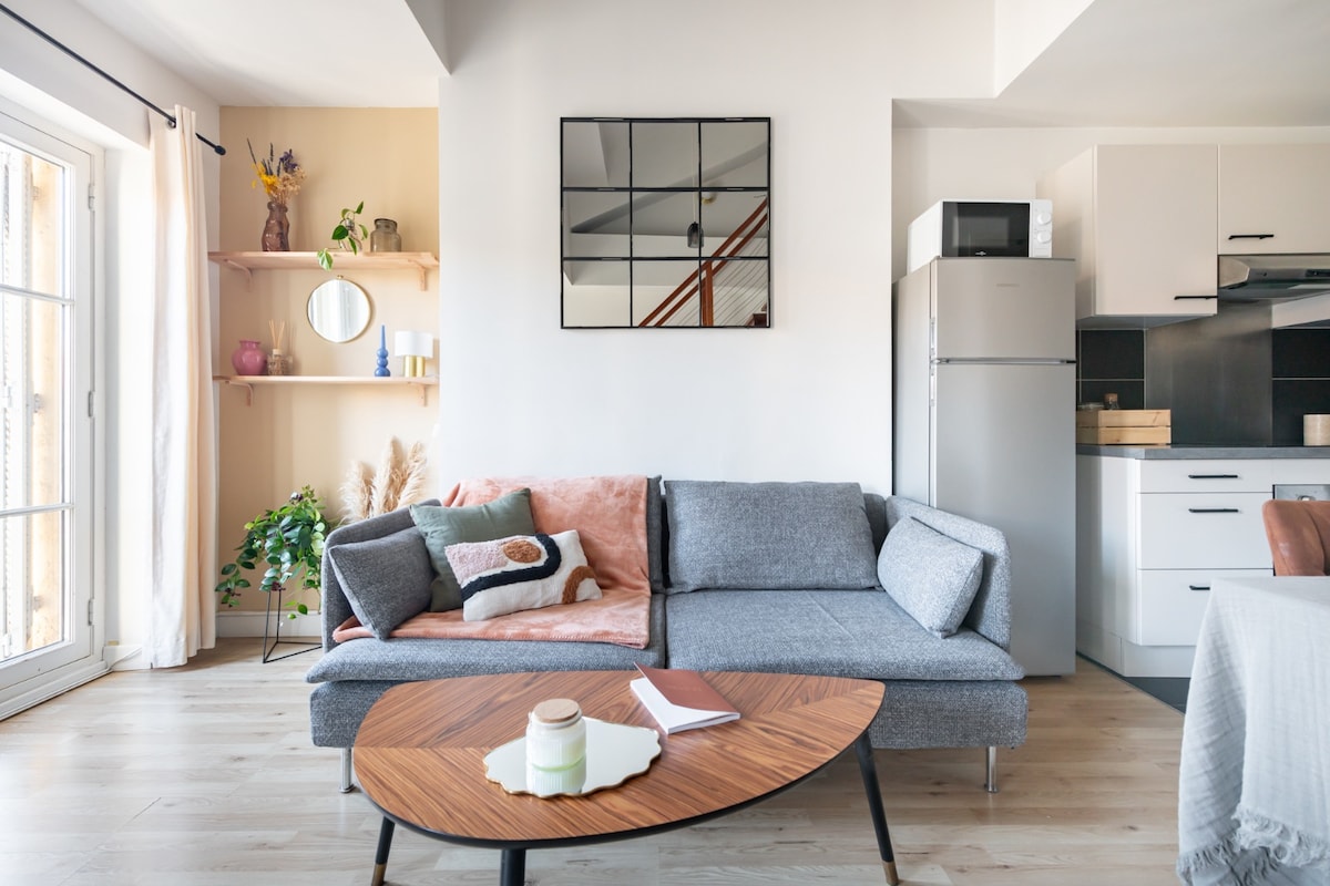 A modern living space features a gray sofa adorned with a pink and brown throw pillow. A round wooden coffee table sits in front, accompanied by a small notebook and a candle. The kitchenette is visible, with a refrigerator and storage units, while a large window lets in natural light.