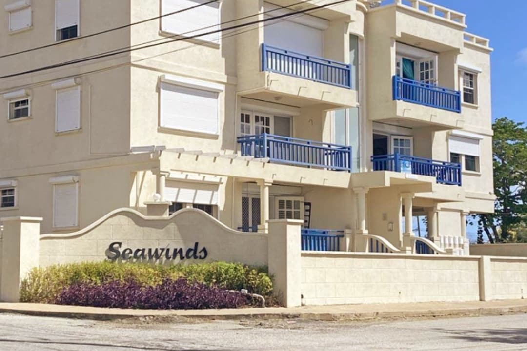 The exterior of a well-maintained building features light-colored walls complemented by blue railings and shutters. The name 'Seawinds' is displayed prominently at the entrance, framed by lush greenery that adds to the welcoming atmosphere.