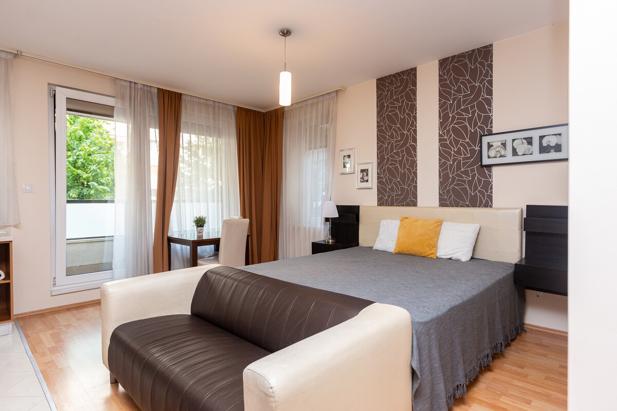 A spacious bedroom is highlighted by a large bed with a grey blanket, flanked by modern nightstands. A small dining table is positioned near a window, enhanced by sheer curtains that allow soft natural light to filter in. Decor includes framed artwork on the wall.