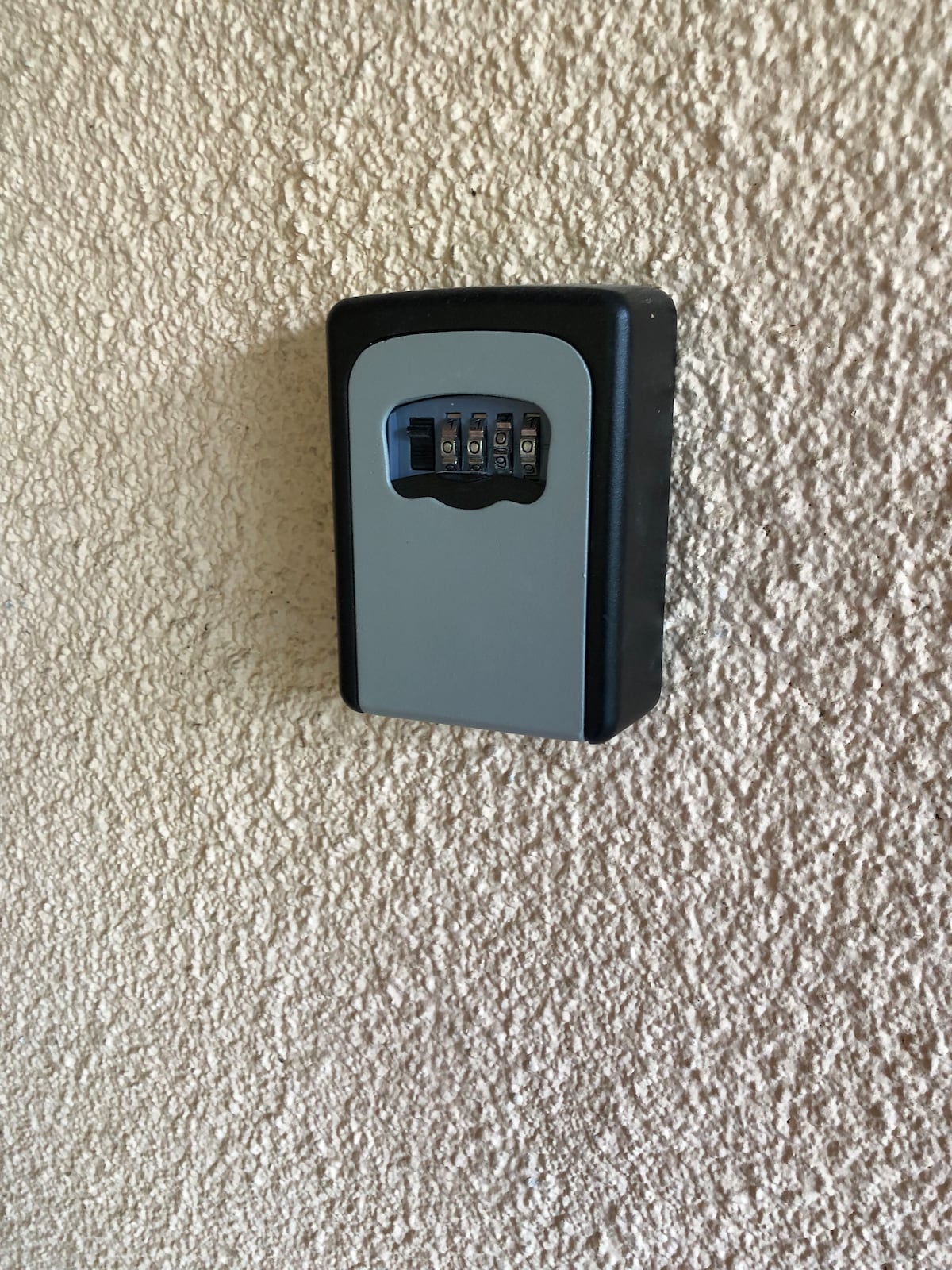A secure keylock box is mounted on a textured wall, featuring a combination dial. The box presents a simple and functional design, allowing access to keys with ease for guests.