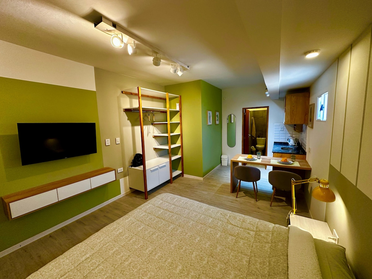 A multifunctional studio space is shown, featuring a spacious bed with a light-colored bedspread. A wall-mounted television enhances entertainment options. A kitchenette area is visible, with wooden cabinets and an adjacent dining setup for two. Soft green walls add a calming feel to the environment.