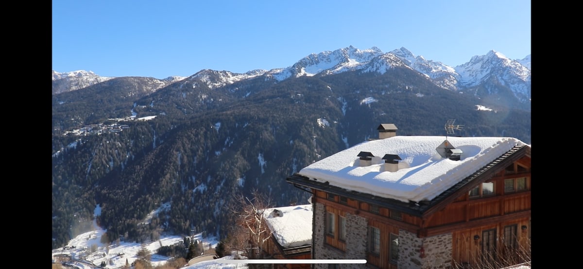 The exterior of a wooden chalet is seen in the foreground, featuring a snow-covered roof. In the background, majestic mountain peaks are highlighted against a clear blue sky, showcasing the natural beauty of the surrounding landscape.