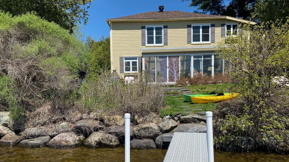 A charming two-story house with yellow siding is set against a natural backdrop. A grassy yard is visible with a small dock leading to the water. A kayak is placed on the grass, enhancing the outdoor atmosphere. Surrounding shrubbery adds greenery to the scene.