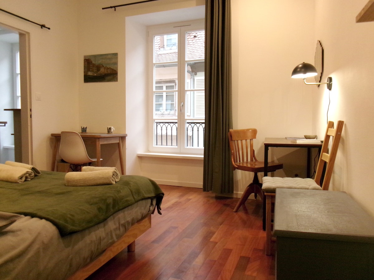 A bright room features a spacious bed with soft linens and towels neatly arranged. A wooden chair and a small desk provide a work area, while a cozy bench sits at the foot of the bed. Large windows allow natural light to fill the room.