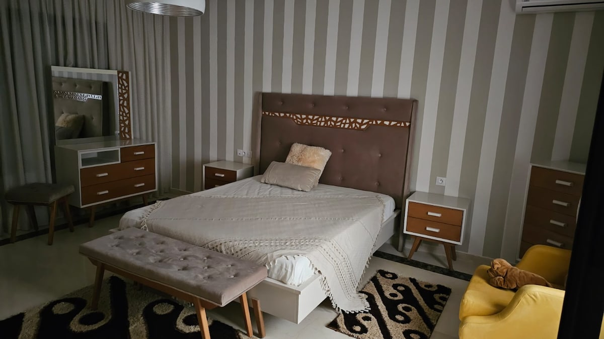 A stylish bedroom features a large, upholstered bed with a neutral-toned blanket draped over it. Complementary nightstands are positioned on either side, and a seating area is provided by a yellow chair and a bench. Soft lighting filters through elegant curtains.