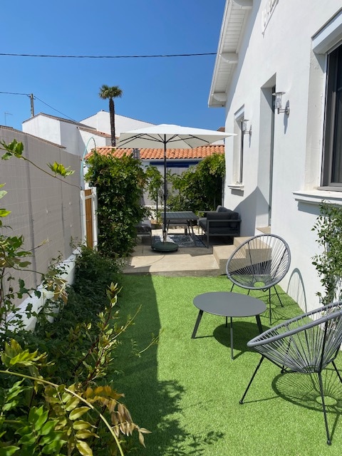 A lush, green outdoor space features artificial turf bordered by shrubs and plants. A seating area includes a sofa and two chairs, accompanied by a small table. An umbrella provides shade over the seating, and a clear blue sky is visible overhead.