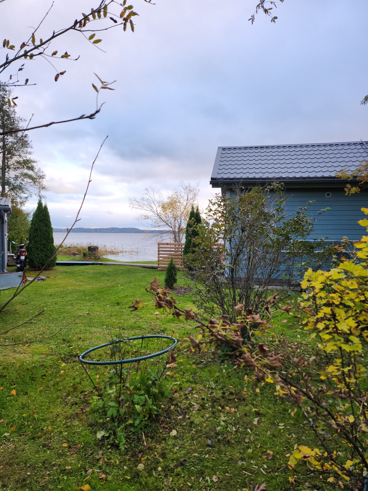 A serene view of a garden area featuring green grass and flowering shrubs. The calm lake is visible in the background, bordered by trees. A wooden fence and a structure with a dark roof are situated to the right, adding to the peaceful landscape.