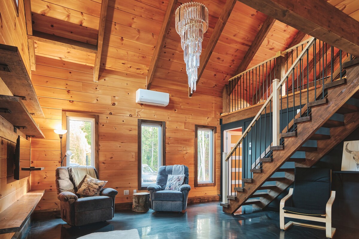 A spacious living area features warm wooden walls and a high ceiling with exposed beams. A chandelier hangs from the ceiling, providing soft light. Comfortable seating is arranged near large windows, and a staircase leads to the upper level.