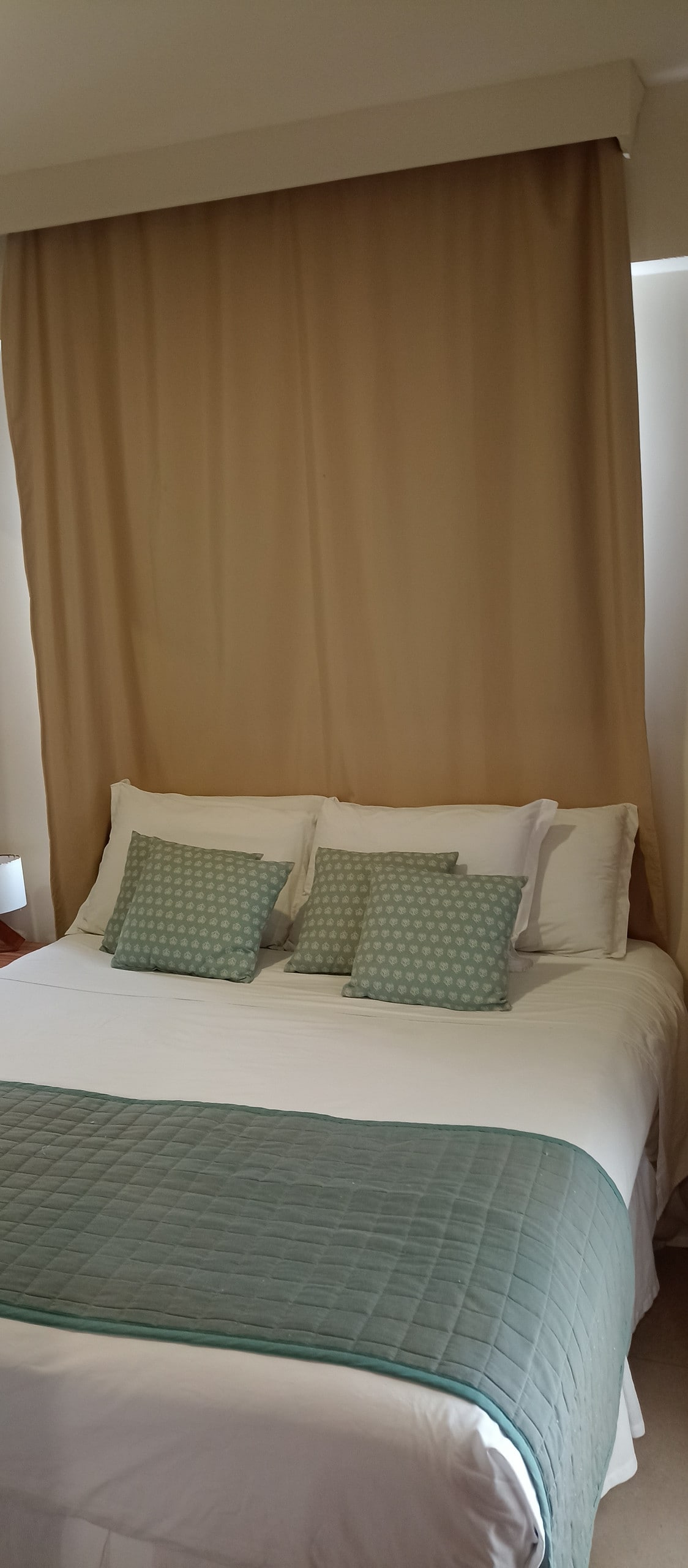 A cozy sleeping area features a spacious bed dressed in white linens, complemented by a light green quilt. Three decorative pillows in coordinating patterns are arranged at the head of the bed, while soft curtains in warm tones frame the space.