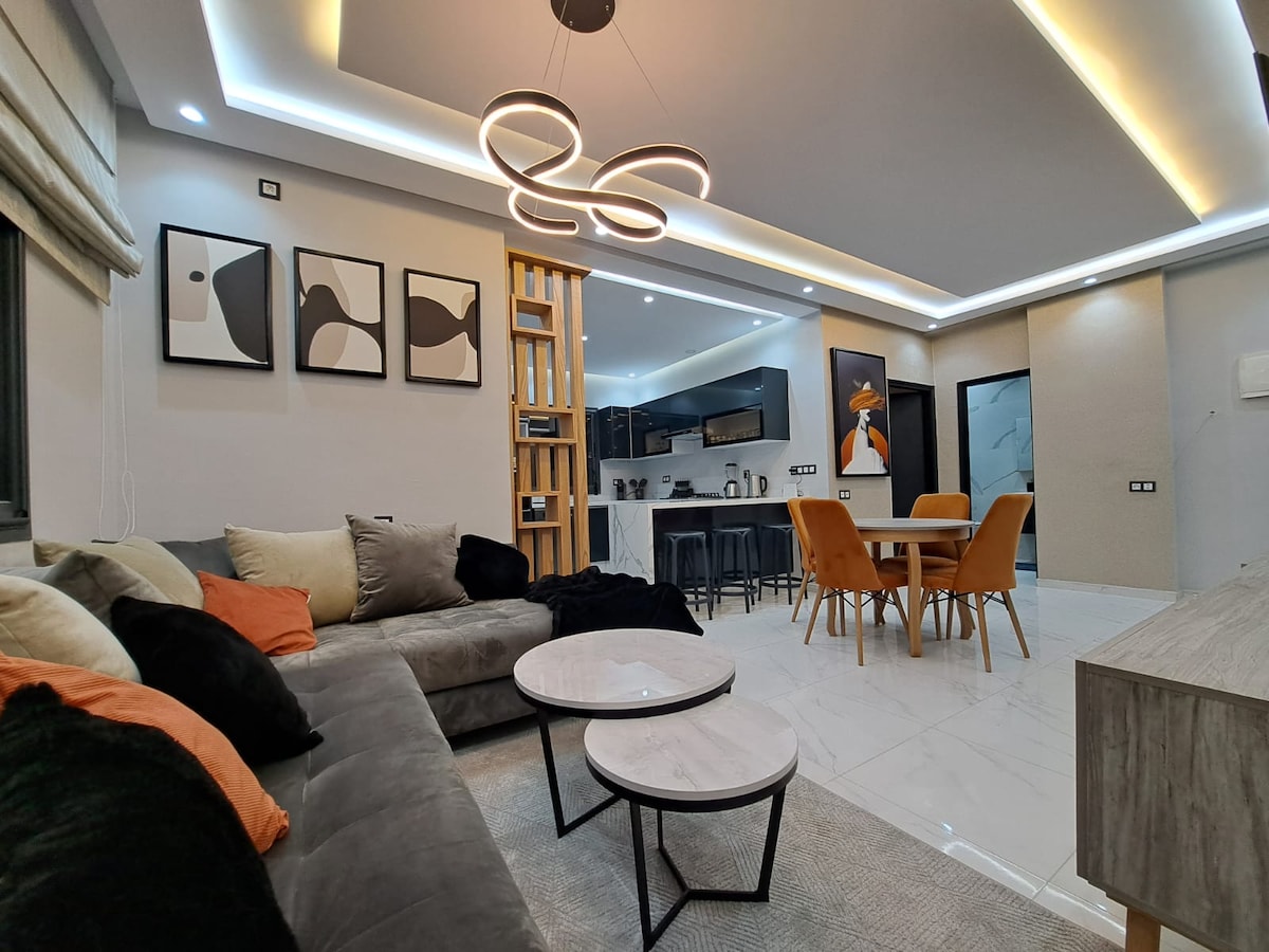 A modern living space features a sectional sofa adorned with various textured cushions, complemented by two round coffee tables. The area is illuminated by a contemporary light fixture, highlighting the open layout with a dining table set for four and stylish artwork on the walls.