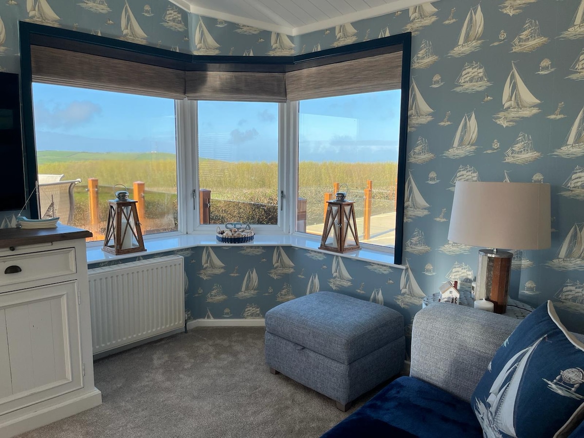 A cozy corner area features a bay window adorned with nautical-themed wallpaper and offers a view of the surrounding landscape. A comfortable ottoman sits nearby, along with a lamp on a white cabinet and decorative lanterns on the windowsill.
