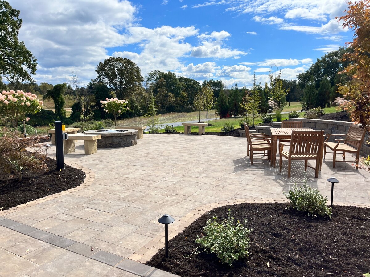 A spacious outdoor patio area features stone pavers, a firepit, and a wooden dining set. Landscaping includes low shrubs and trees, with pathways guiding the view to the surrounding greenery under a clear sky.