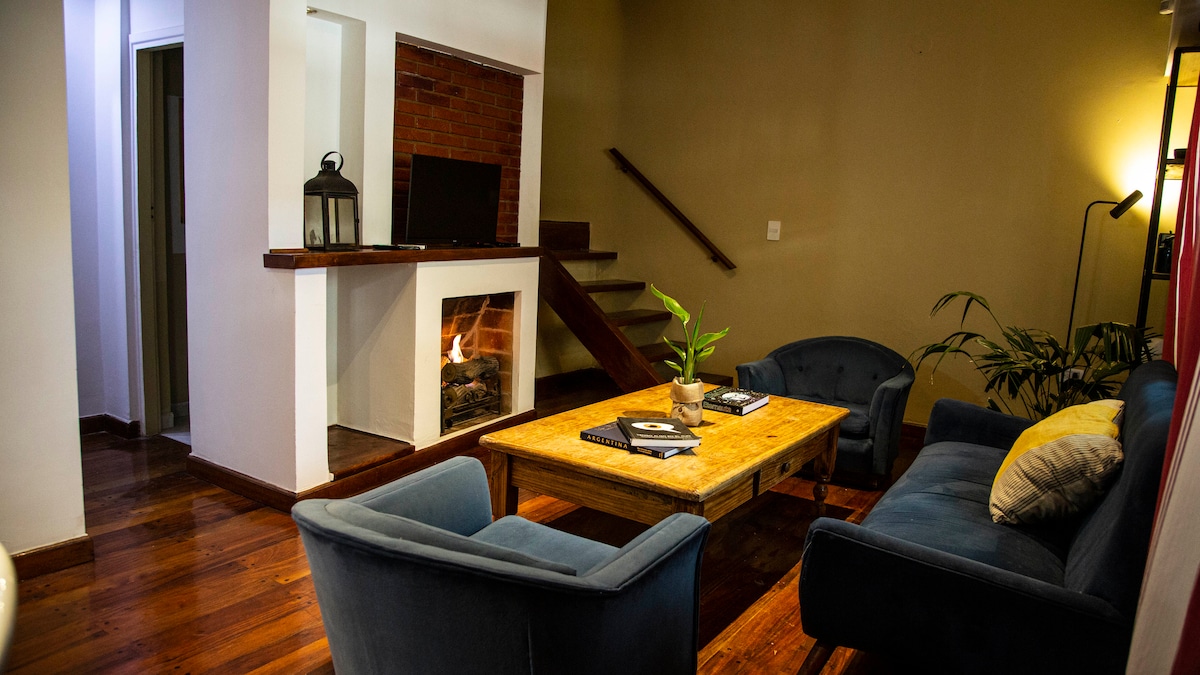 A welcoming living area features a wooden table at its center, surrounded by plush blue armchairs and a comfortable sofa. A gas fireplace adds warmth, while a flat-screen TV is positioned on a shelf nearby. Light wood flooring and a staircase enhance the space's open feel.