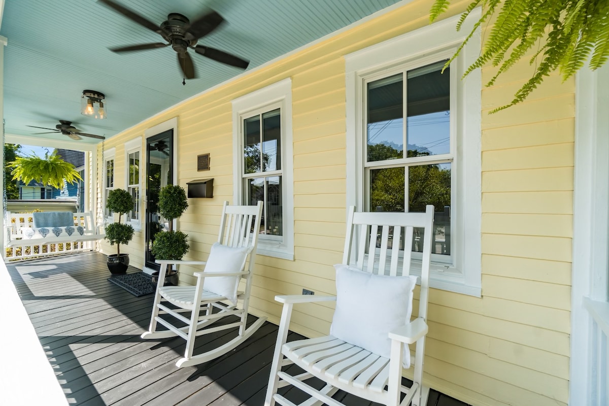 A charming front porch features two white rocking chairs with plush cushions, positioned on a dark wood floor. Large windows are framed in white, allowing natural light to illuminate the yellow exterior walls. Ceiling fans are installed for added comfort.