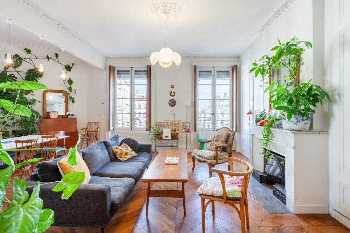 A spacious living area features large windows allowing natural light, adorned with plants along the walls. A comfortable seating arrangement includes a dark sofa and wooden chairs around a coffee table. Decorative elements include framed art on the walls and a fireplace with plants nearby.