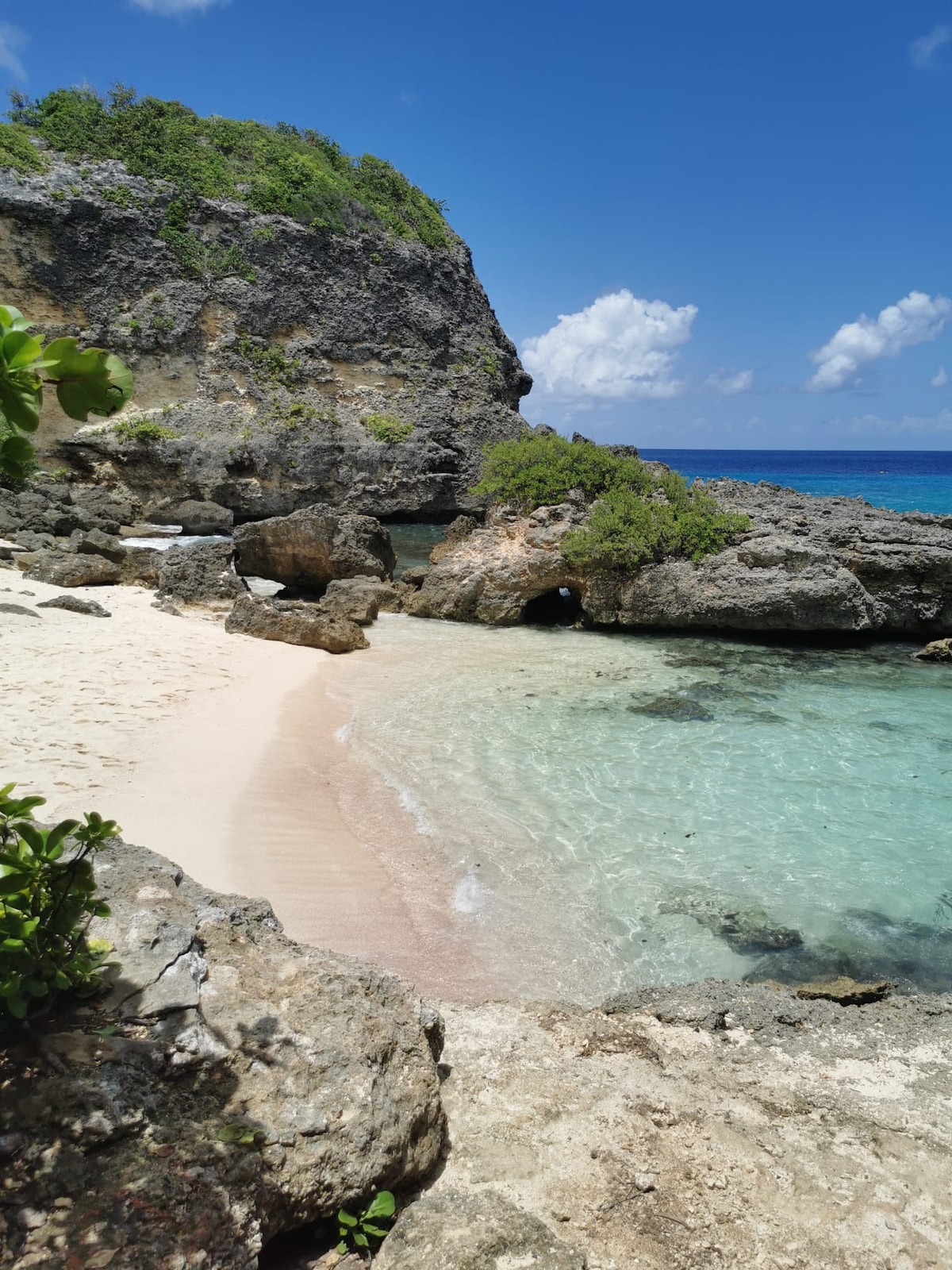 A secluded beach is framed by rocky cliffs and lush greenery. The soft sand transitions to clear turquoise water with gentle waves lapping at the shore. Sunshine illuminates the scene, highlighting the natural beauty of the coastal landscape.