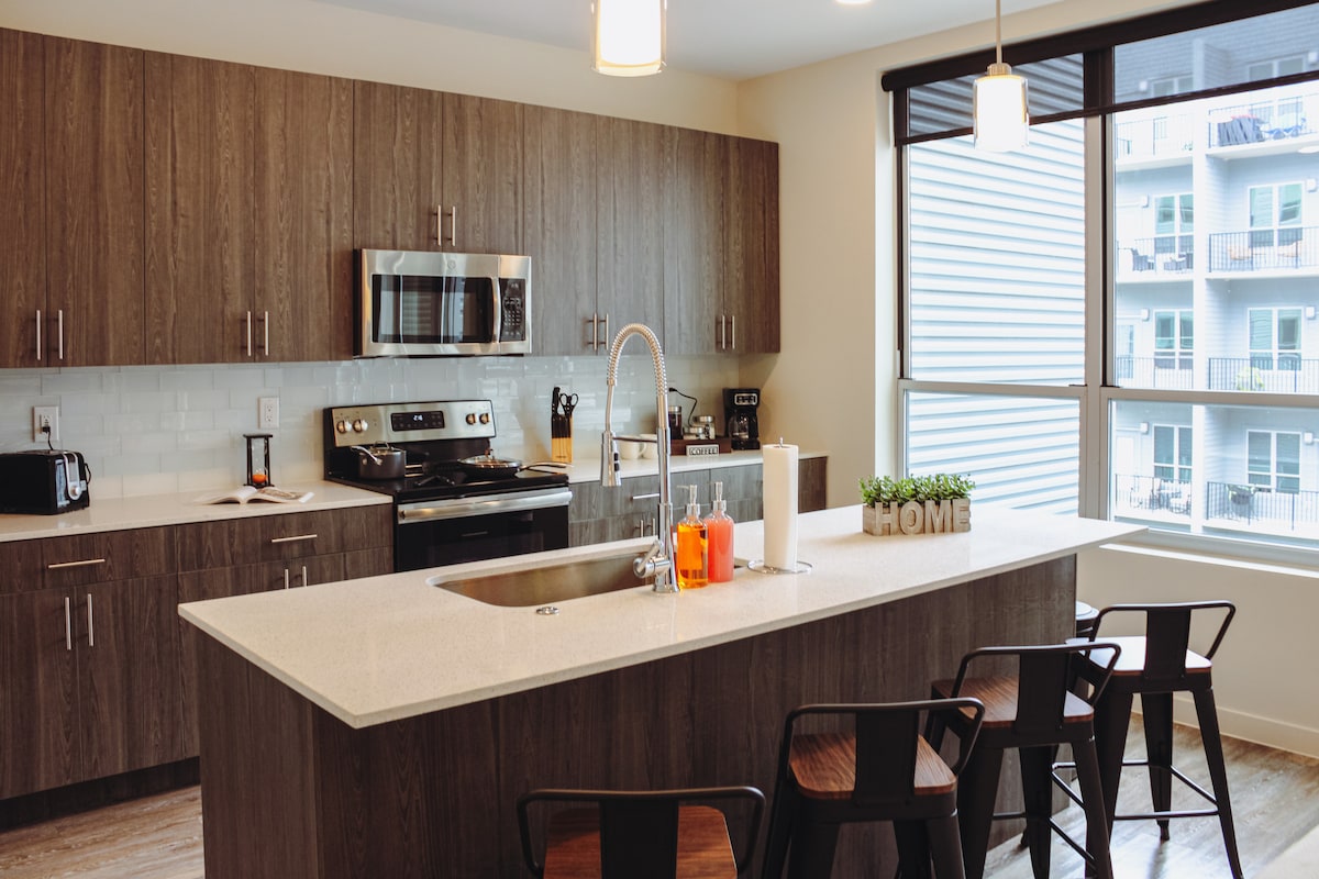 The modern kitchen features sleek wooden cabinetry and stainless-steel appliances. A spacious island with seating for four is highlighted, while natural light pours in through large windows. Countertops are neat and uncluttered, showcasing a cozy decoration that spells 'HOME.'