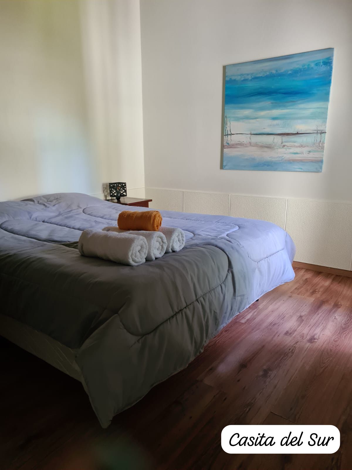 A sleeping area features a spacious bed covered with a light gray comforter. Rolled towels are neatly arranged on the bed, complemented by a warm-toned blanket. A colorful landscape painting hangs on the wall, while natural light filters softly through the room.