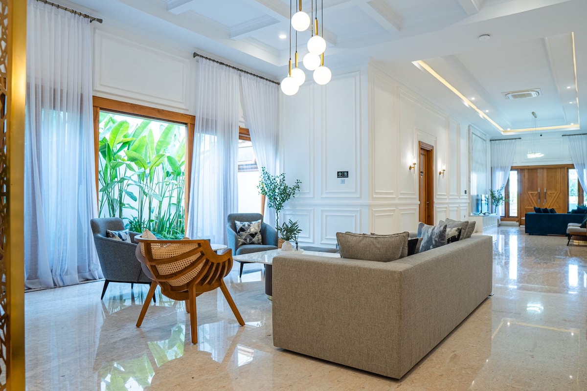 A spacious living area features natural light streaming through large windows, framed by sheer curtains. Comfortable seating includes a gray sofa and two armchairs, arranged around a unique wooden table. Lush greenery is visible outside, enhancing the inviting ambiance.