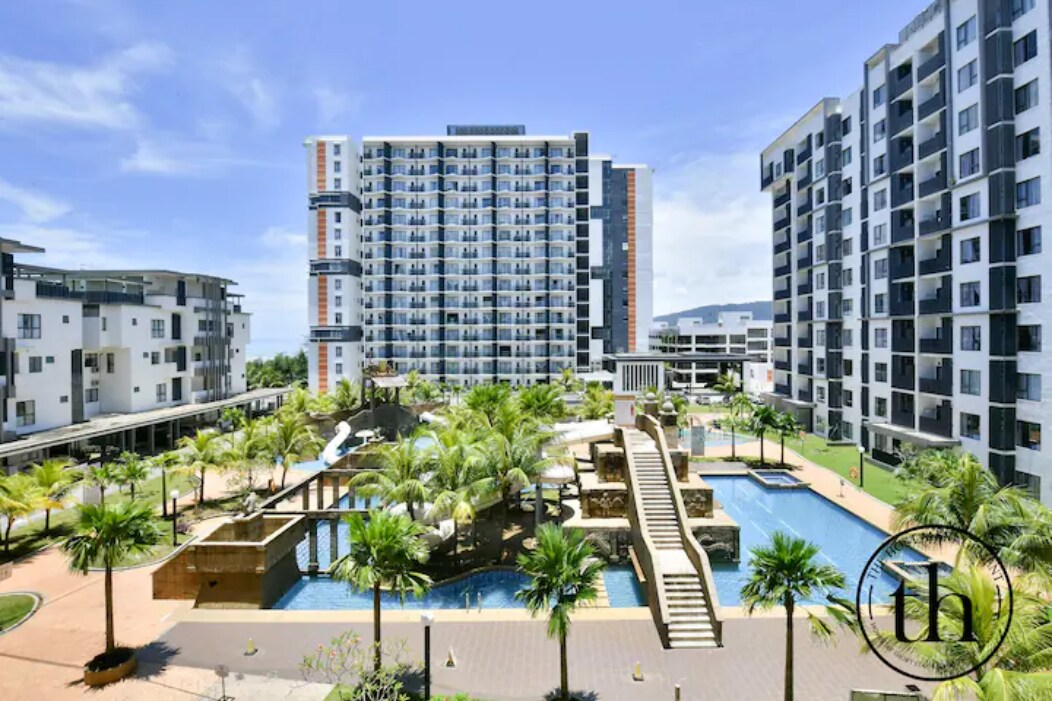 The image captures a resort-style apartment complex surrounded by lush greenery. Multiple palm trees are visible, framing a central swimming pool area with a water theme park. The architecture features modern buildings, complementing the vibrant outdoor setting.