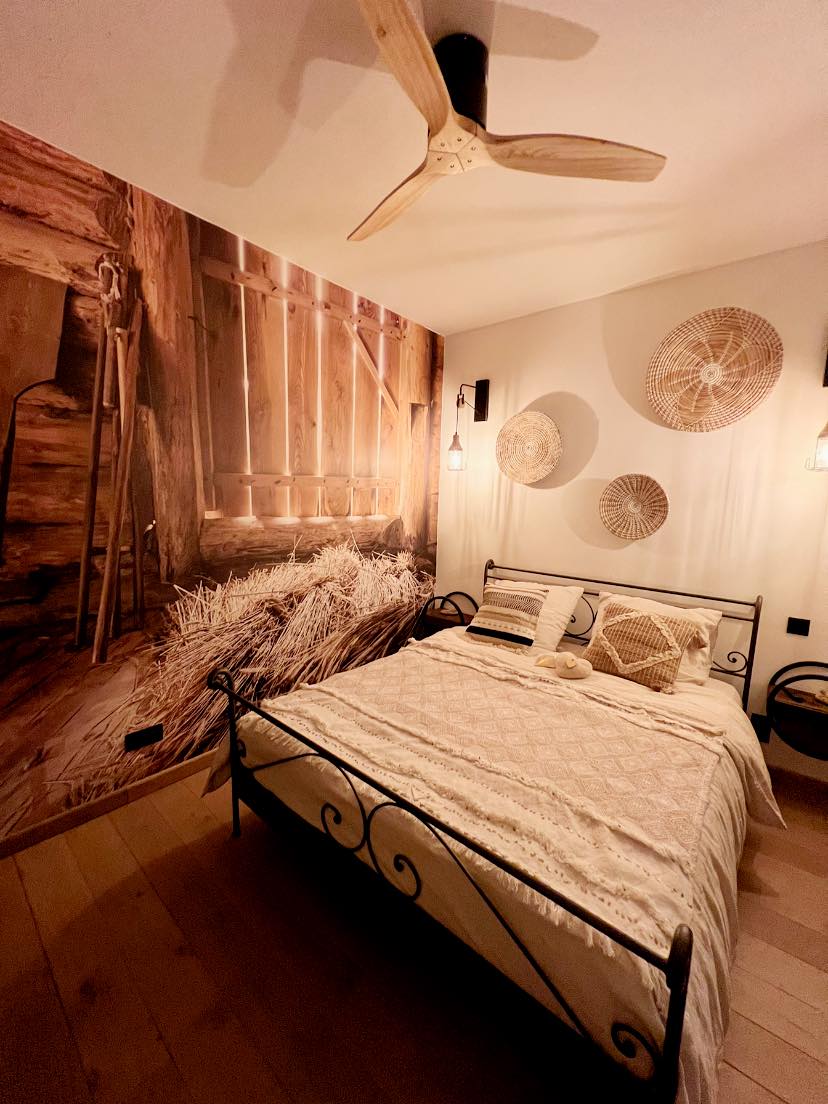 The sleeping area features a double bed with light bedding, surrounded by a rustic mural depicting a barn interior. Wall decorations include woven circular art pieces. Soft lighting is provided by fixtures beside the bed, while a ceiling fan adds comfort.