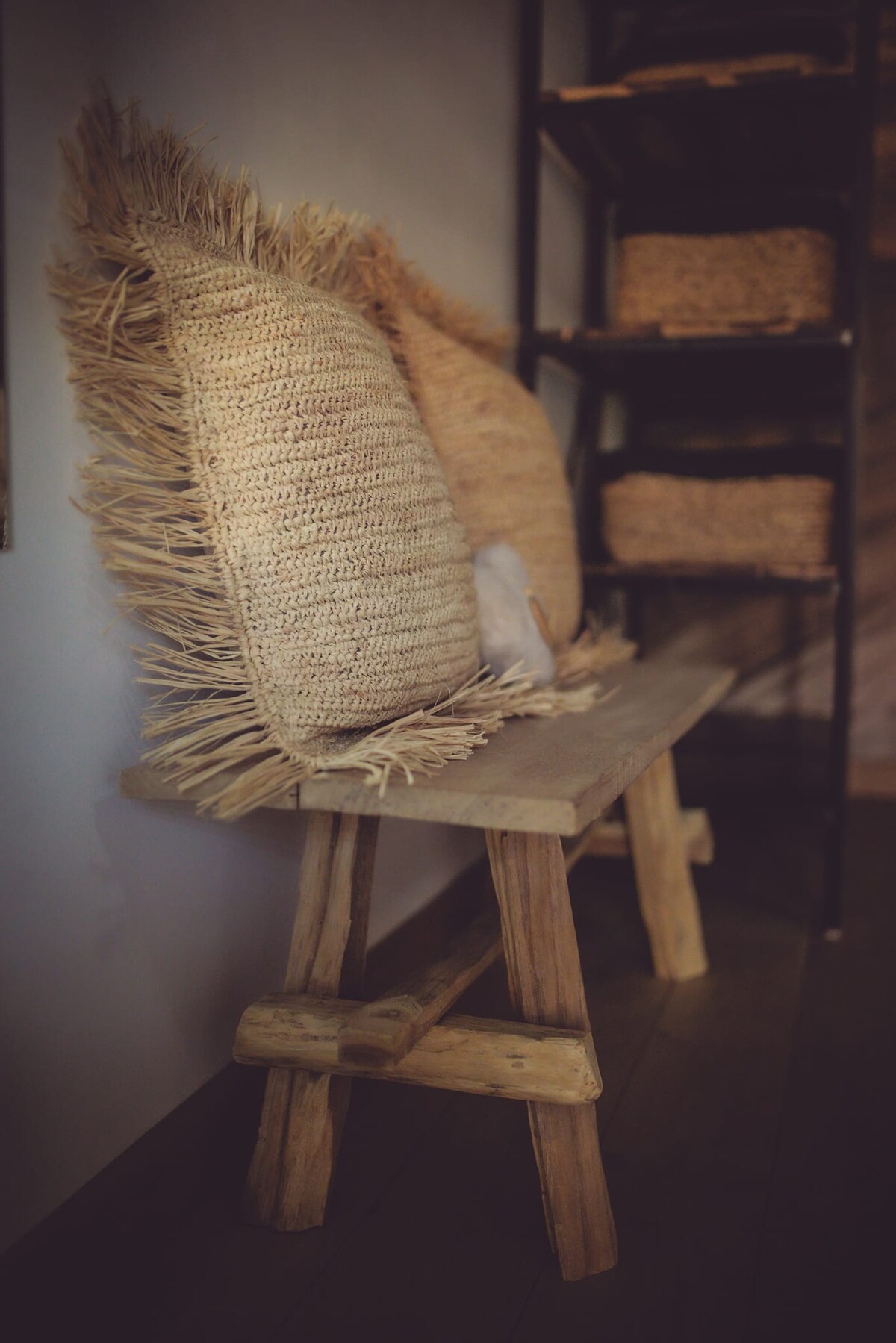 A wooden bench is positioned against a wall, adorned with two textured throw pillows featuring fringed edges. A shelf with woven baskets is visible in the background, contributing to a cozy and organized setting.