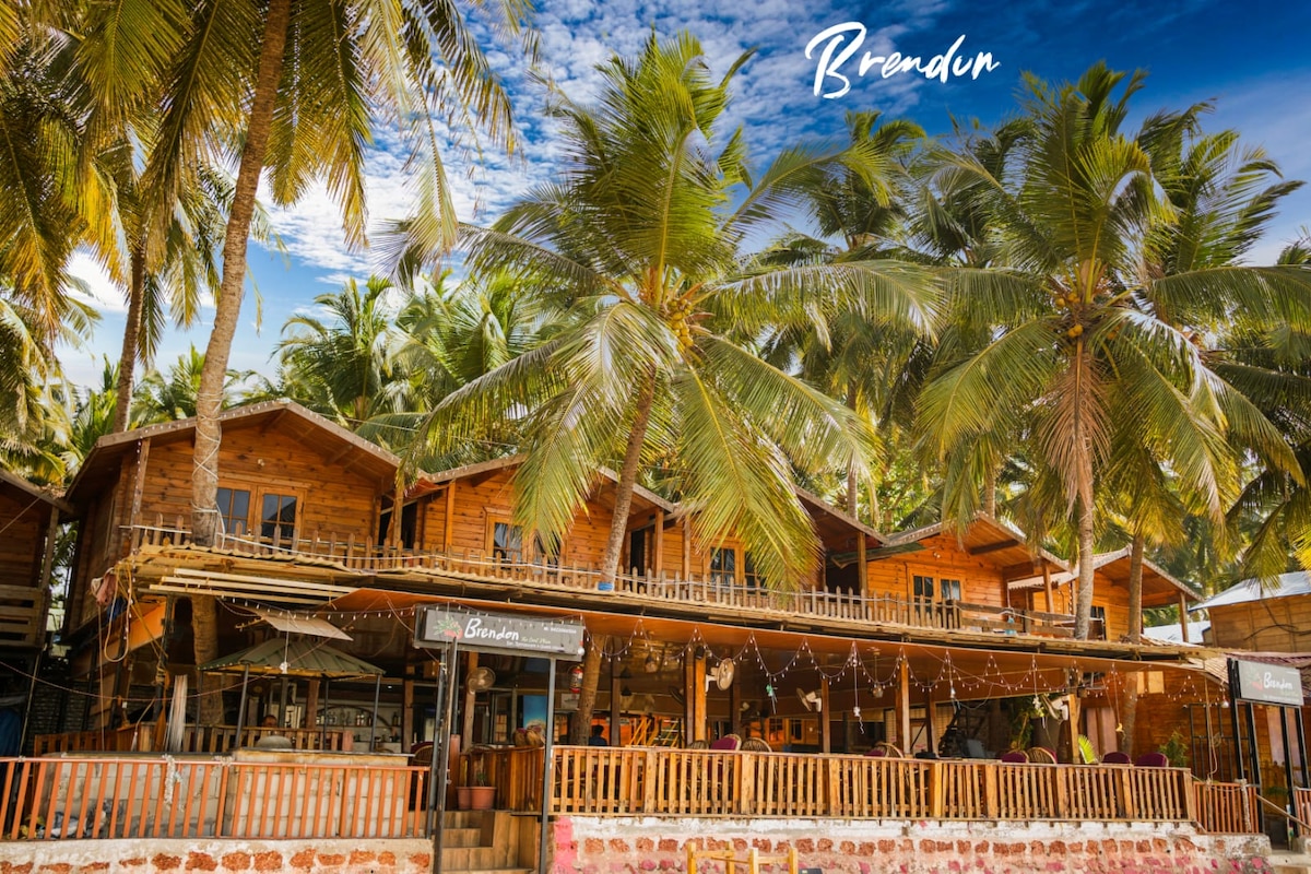 A charming multi-level wooden structure is surrounded by tall palm trees, offering a relaxed tropical atmosphere. The building features a spacious balcony with railing and string lights, while large windows provide views of the vibrant outdoor scenery.
