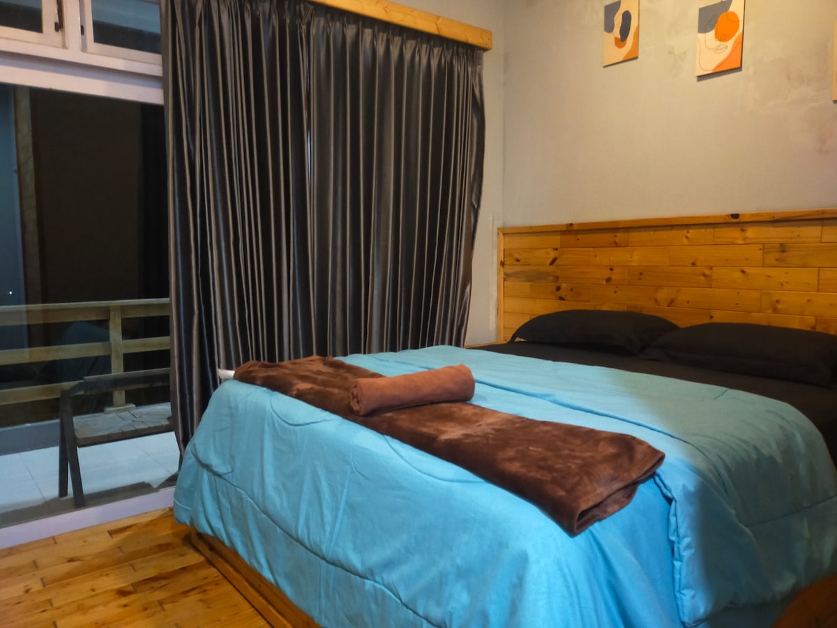 A spacious bed is dressed with a light blue cover and dark brown blankets. The wall features wooden panels, and large curtains frame the window. Two pieces of wall art are displayed above the bedding, contributing to the room's aesthetic. A balcony is visible through the window.